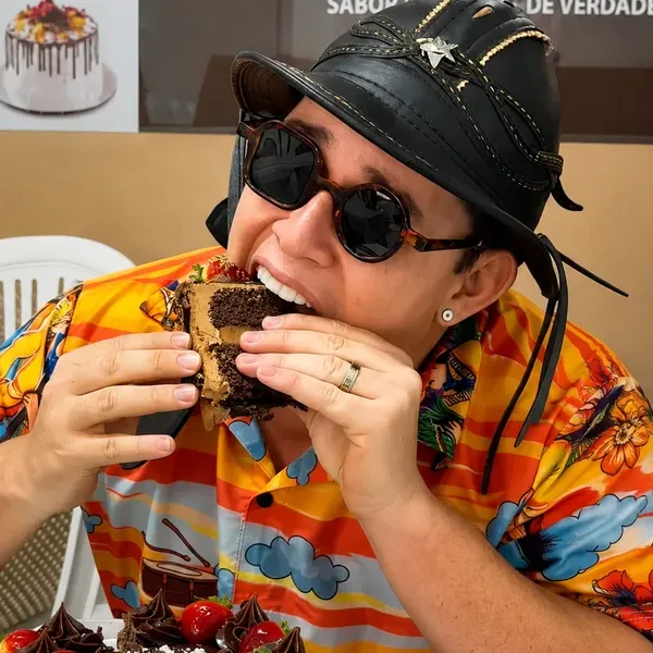 Chaveirinho de óculos escuros mordendo uma fatia de bolo de chocolate em uma confeitaria